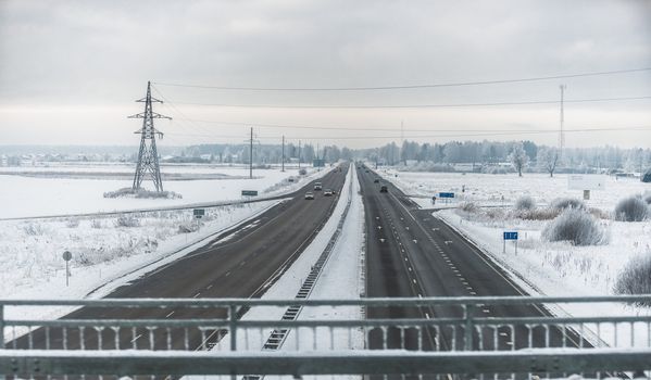Panorama of a highway with snow in countryside in winter. A few cars are visible in a distant horizon. A view from a bridge.