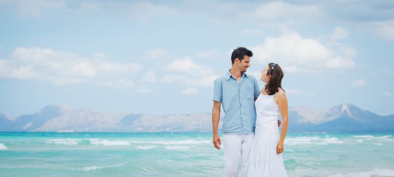 Happy young man and woman couple walking, laughing and holding hands on a deserted tropical beach with bright clear blue sky