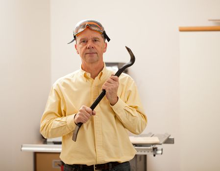 Senior male in a home workshop facing the camera and holding a crowbar