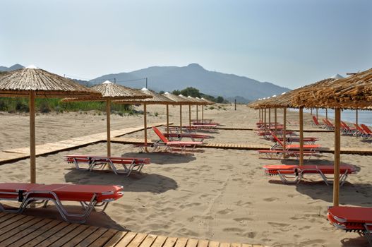Straw umbrellas and rows of lounge sunbed chairs at sandy beach in Greece.