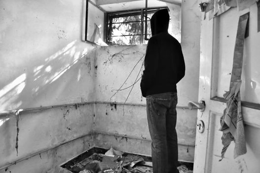 Hooded male figure looking through window of abandoned house. Black and white.