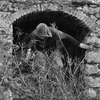 Hooded man obscured by overgrown plants under arched recess of rural ruin.