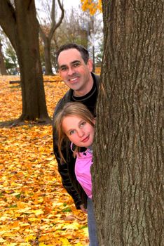 Father and daughter peaking from a tree trunk in autumn park