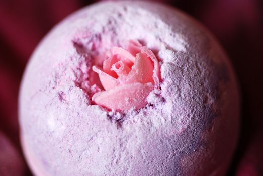 A close-up photograph of a luxurious bath product, with an embedded delicate pink rose, on a deep red velvet background