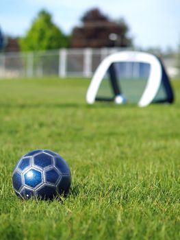 A blue soccer ball on a field of green grass on a sunny day with the goal in the background.