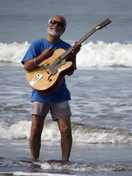 A senior Indian rockstar playing an acoustic guitar in beach waters.                               