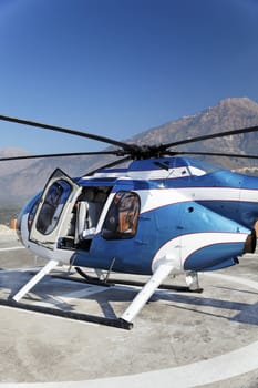 Abstract portrait of small metallic blue and white helicopter stationed on a helipad with open door, blue skies with negative spaces