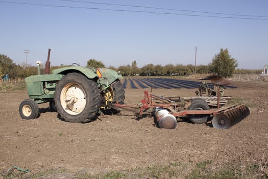 a farm tractor with a plow on the back with farm rows in the background.
