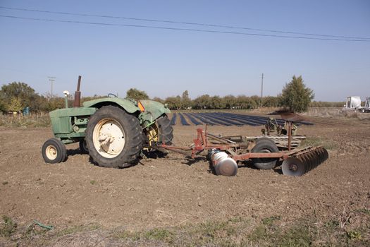 a farm tractor with a plow on the back with farm rows in the background.