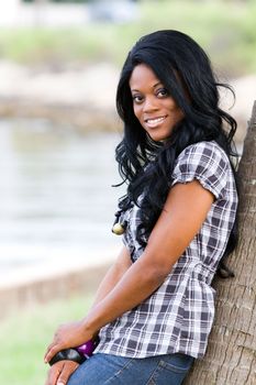 Casually dressed happy young African American black woman leans against a tree smiling.