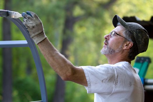 Mature man paints ironwork with paintbrush applying primer to the metal.