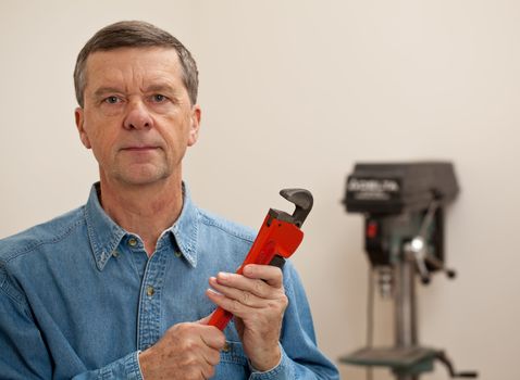 Senior male in a home workshop facing the camera and holding a large wrench