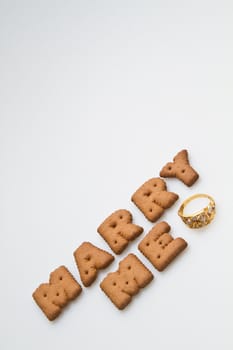 Angled view of the words marry me arranged by brown biscuits with gold ring on white background portrait orrientation