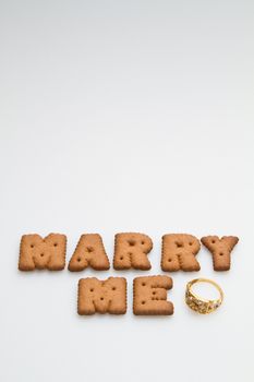 Marry me wording from brown biscuits with gold ring on white surface portrait orientation