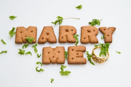 Marry me wording by brown biscuits with green leaves and gold ring on white surface in landscape orientation