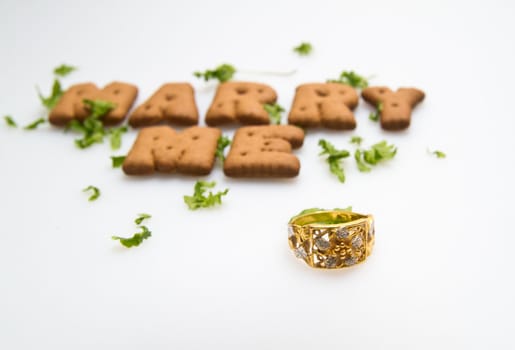 Marry me wording by brown biscuits with green leaves and gold ring on white surface in landscape orientation