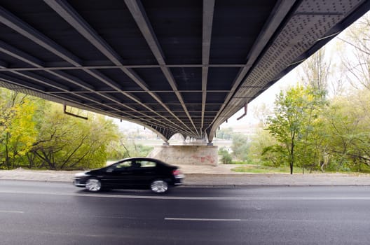 Bridge underneath over the river and road and black car going fast out of focus.