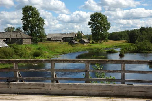View to northern russian village by the river from wooden bridge, clouds in sky