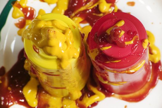 Messy mustard and ketchup squeeze bottles standing up on a plate.