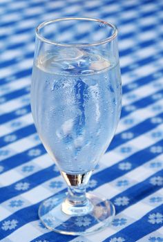 A glass of iced water, standing on a blue and white tablecloth, ice cubes in glass and condensed water on glass due to heat