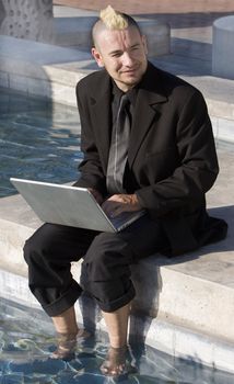 Businessman with a punk haircut works on a laptop computer with his feet in a fountain.