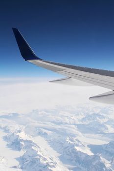 wing of a passenger jet at high altitude with snow covered mountain terrain in the mist below 