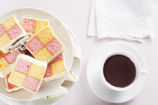Battenberg cake on a two tiered serving plate and a cup of tea on the table