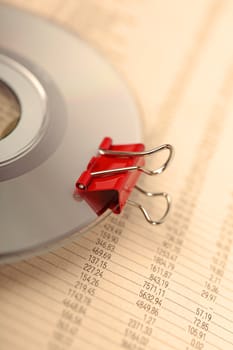 Macro of red paperclip attached to compact disk, laying on newspaper with financial data.