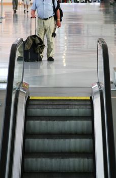 a bodyshot of a man standing by escalators in an airport with hand luggage