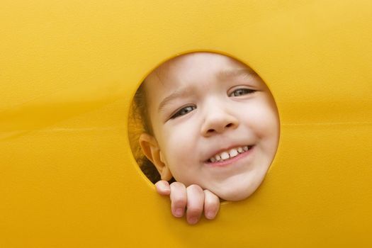 A little girl sticks her face through a hole in a yellow plastic piece of playground equipment