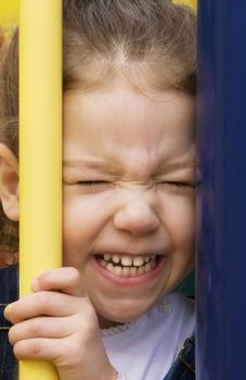 Little girl on play equipment scrinches her face between two bars.