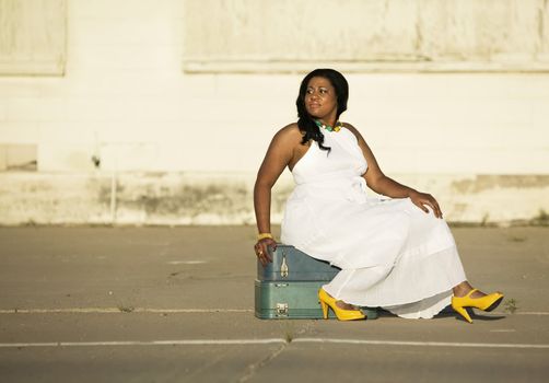 African American woman waits by the side of the road sitting on two suitcases.