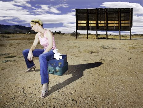 Woman sitting on suitcases in front of an old billboard waiting in the desert.