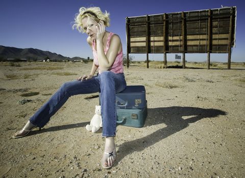 Woman sitting on suitcases in front of an old billboard waiting in the desert.