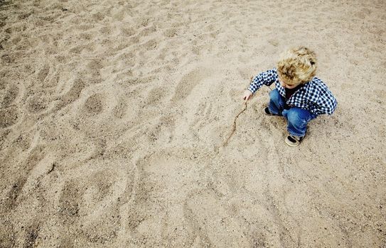 Little boy with a stick plays in a large patch of rough sand.