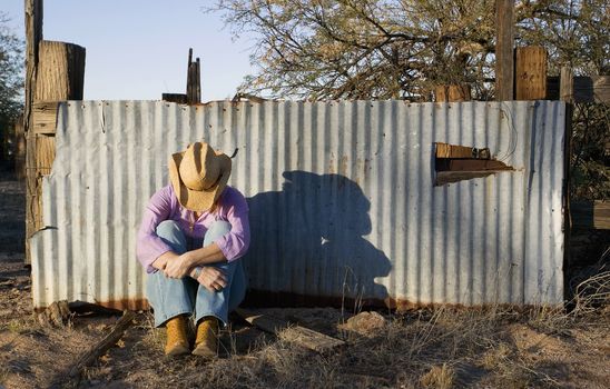Woman wearing a straw cowboy hat with her head down leaning against a corrugated metal fence.