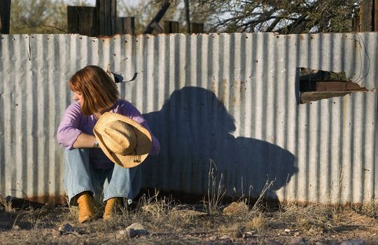 Woman in a Cowboy hat sitting against a Corrugated Metal fence and Looking to her Right.