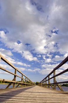 Long wooden bridge over the lake with handrail made of planks and blue cloudy sky.