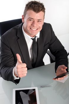 Enthusiastic young businessman showing thumb up while using a smart phone at his desk
