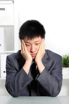 Despondent Asian businessman sitting at his desk with his chin resting in his hands staring morosely at the desktop
