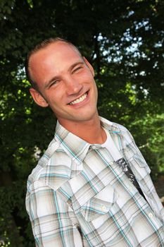 Smiling sexy handsome young man in a casual shirt standing outdoors in front of greenery