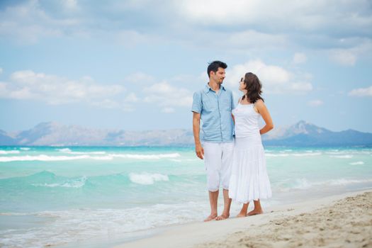 Happy young man and woman couple walking, laughing and holding hands on a deserted tropical beach with bright clear blue sky