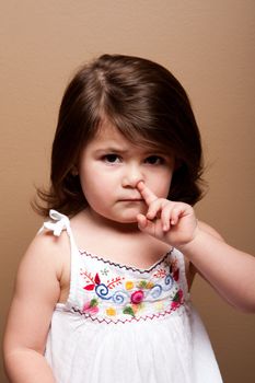 Cute mischievous toddler girl with finger in her nose fishing for boogers snot, on brown.