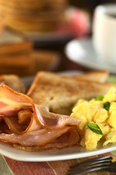 Fried bacon and scrambled eggs with toast bread, pancakes and coffee in the back (Selective Focus, Focus one third into the bacon and on the front of the eggs)