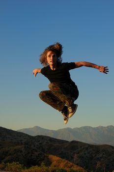 Caucasian male 20-40 in sportswear jumping above mountain peaks, facing forward, arms widespread, knees up.
