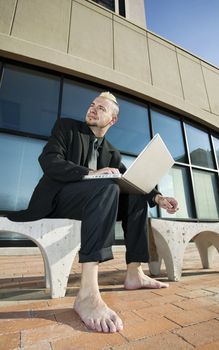 Barefoot businessman with a punk haircut works on his laptop computer on a bench in a business park.