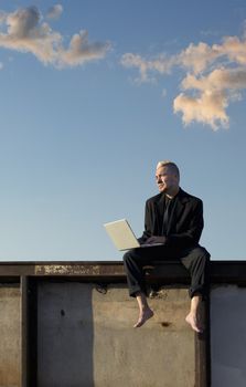 Barefoot businessman with a punk haircut works on his laptop computer sitting on a wall.