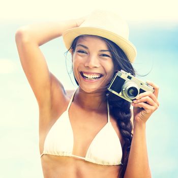 Beach woman with vintage retro camera having fun playful laughing in bikini on blue ocean background wearing beach hat. Multicultural Asian / Caucasian girl.