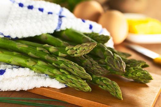 Raw green asparagus wrapped in a dish towel lying on a wooden board with eggs and butter in the back (Selective Focus, Focus on the three asparagus heads in the front)