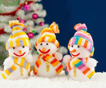 Three snowmen in front of the decorated white evergreen tree over the blue background